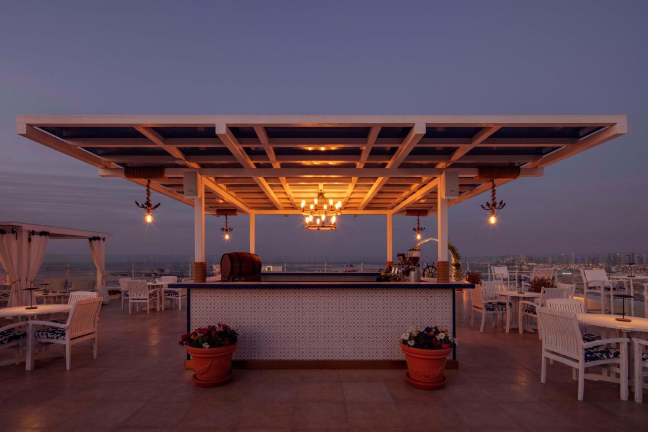 Hotel Hyatt Regency Dubai - Corniche Esterno foto The photo shows a picturesque outdoor bar or dining area situated on a rooftop or terrace. It features a wooden structure with a slatted roof, illuminated by warm lights, including a chandelier and hanging lamps. The bar is adorned with potted plants